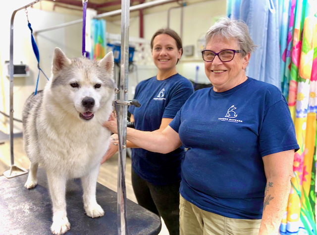 Two TopDog staff members smiling beside a husky standing on the grooming table.